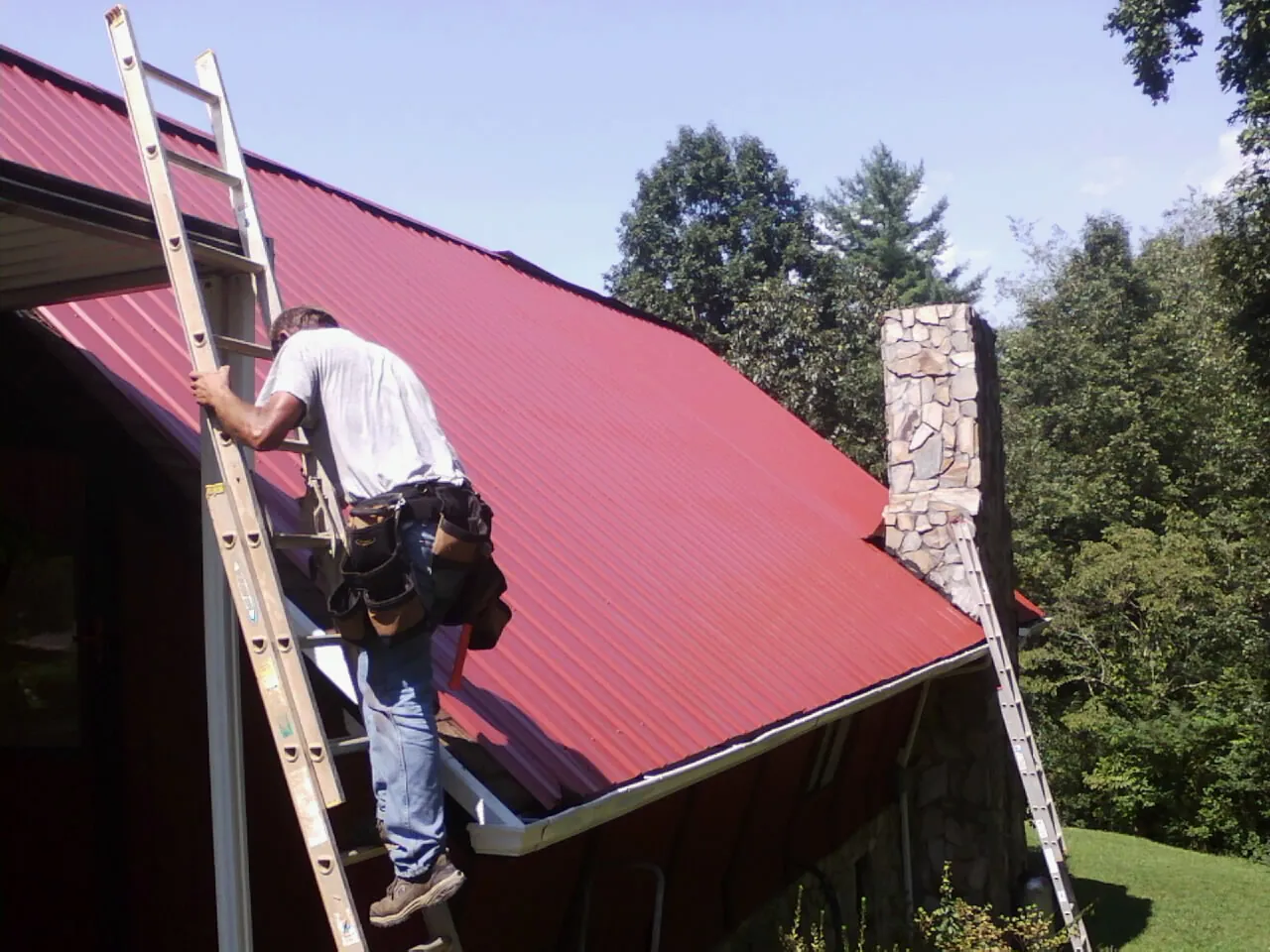 Roofer on a ladder beside a red metal roof and stone chimney — Soffit & Fascia Repair by Roofing Brothers in Grand Coulee